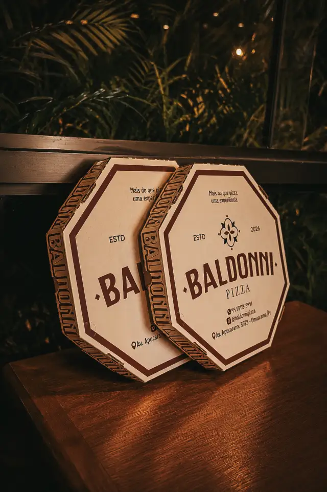 Caixas octogonais de pizza da Baldonni sobre a mesa de madeira, com o brasão da casa e o slogan Mais do que pizza, uma experiência